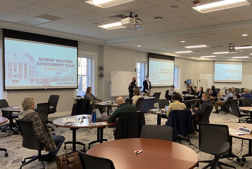 Individuals sit at tables while listening to a speaker and watching a slide presentation titled "winter weather summit."